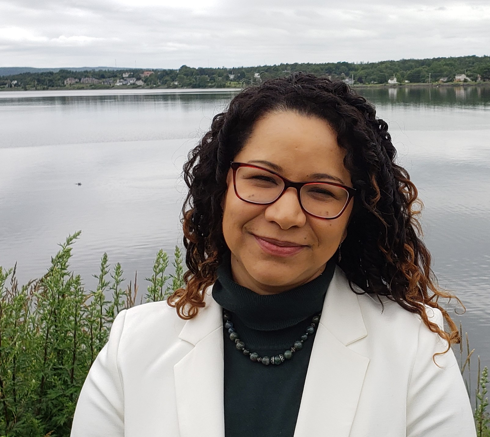 Femme à lunettes et aux longs cheveux noirs bouclés, en blouse de laboratoire, souriant devant un lac. Photo : Dre Lilian Navarro