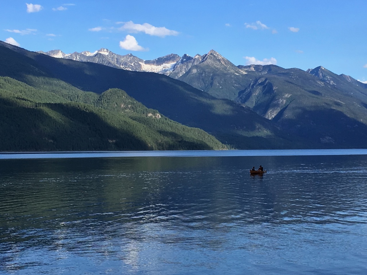 Un lac avec deux personnes dans un bateau, avec des montagnes boisées derrière, et des pics enneigés, avec un ciel bleu.